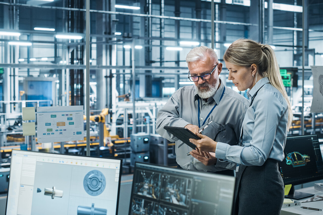 Two specialists are analysing digital production data on a tablet in a modern manufacturing environment to evaluate software solutions and digital use cases.