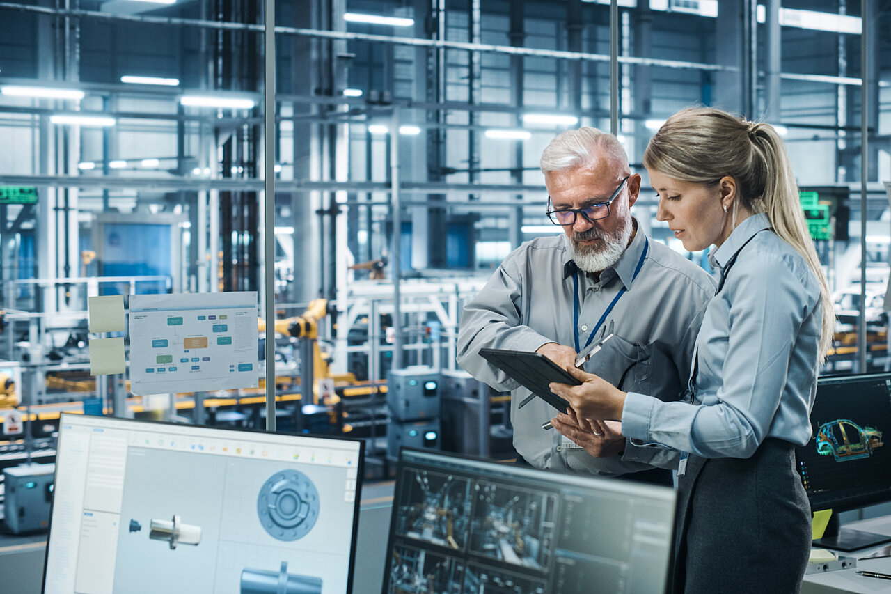 Two specialists are analysing digital production data on a tablet in a modern manufacturing environment to evaluate software solutions and digital use cases.
