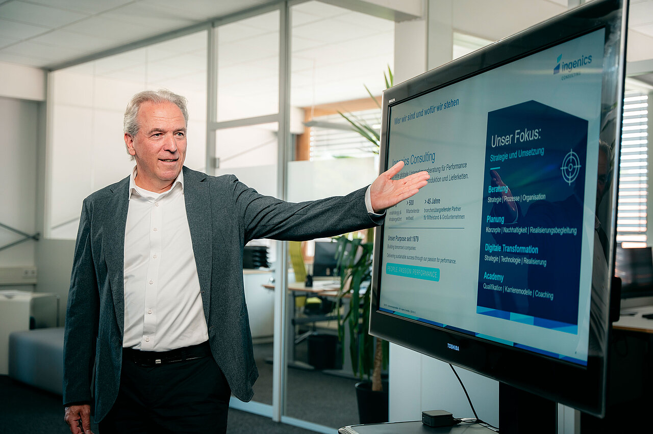 A person is presenting content from Ingenics Consulting on a screen during a meeting in an office setting.